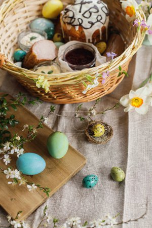 Foto de Natural dyed easter eggs on background of homemade easter bread, ham, beets, butter on rustic table with spring blossoms and linen napkin. Top view. Traditional Easter food in basket - Imagen libre de derechos