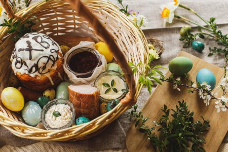 Foto de Traditional Easter food in basket. Homemade easter bread, natural dyed easter eggs, ham, beets, butter, cheese on rustic table with spring blossoms and linen napkin. Top view - Imagen libre de derechos