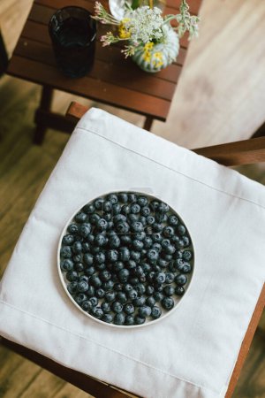 Photo for Fresh blueberries on modern ceramic plate and wildflowers bouquet in rustic room. Summertime in countryside. Healthy food aesthetics. Summer berries top view - Royalty Free Image