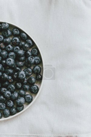 Photo for Blueberries in modern ceramic plate flat lay. Summertime in countryside. Healthy food aesthetics. Summer berries on soft linen background, moody banner - Royalty Free Image