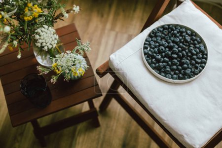 Photo for Fresh blueberries on modern ceramic plate and wildflowers bouquet in rustic room. Summertime in countryside. Healthy food aesthetics. Summer berries - Royalty Free Image