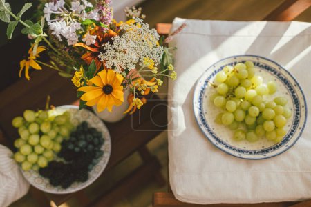 Téléchargez les photos : Summertime in countryside. Wildflowers bouquet on background of fresh blueberries, blackberries and grapes in sunlight in rustic room. Summer flowers and berries in light, home aesthetics. - en image libre de droit