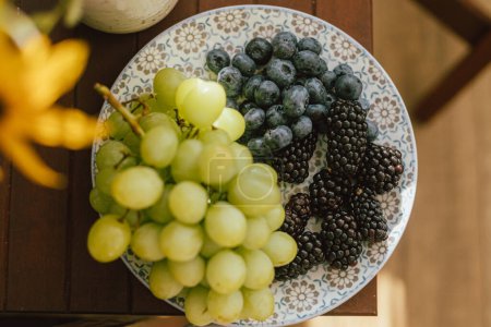 Téléchargez les photos : Bleuets frais, mûres et raisins au soleil sur plaque en céramique dans une pièce rustique. l'esthétique des aliments sains. Baies d'été en vue dégagée. L'été à la campagne - en image libre de droit