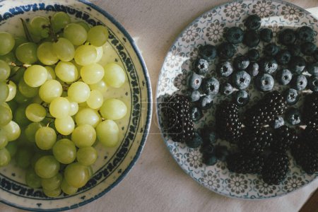 Téléchargez les photos : Fresh blueberries, blackberries and grapes in sunlight on ceramic plate flat lay. Healthy food aesthetics. Summer berries in light on soft linen background. Moody banner - en image libre de droit