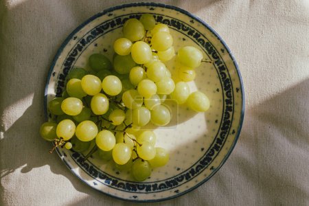 Photo for Fresh grapes in sunlight on ceramic plate flat lay. Healthy food aesthetics. Summer fruits in light on soft linen background. Summertime in countryside, moody banner - Royalty Free Image