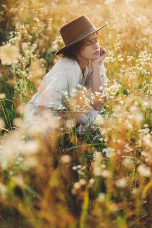 Foto de Elegante mujer boho en sombrero sentado entre flores silvestres en la cálida luz del atardecer. Delicias de verano y viajes. Joven hembra en tela de lino rústico relajándose en el prado de verano. Momento atmosférico - Imagen libre de derechos