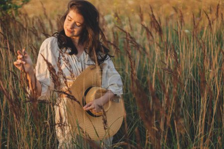 Photo for Stylish boho woman with straw hat posing among wild grasses in evening. Summer delight and travel. Young carefree female in rustic linen cloth relaxing in summer meadow. Atmospheric moment - Royalty Free Image