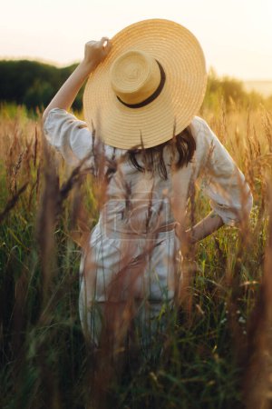 Foto de Elegante mujer boho con sombrero de paja posando entre hierbas silvestres en la luz del atardecer, vista trasera. Verano deleite y tranquilidad. Joven mujer despreocupada en tela de lino rústico relajante en el prado de verano - Imagen libre de derechos