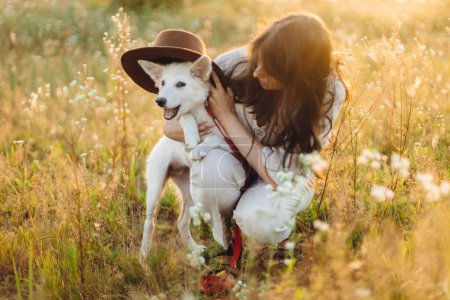 Photo for Stylish happy woman playing with cute dog with hat among wildflowers in sunset light. Summer vacation with pet. Young carefree female relaxing with white danish spitz in summer meadow - Royalty Free Image