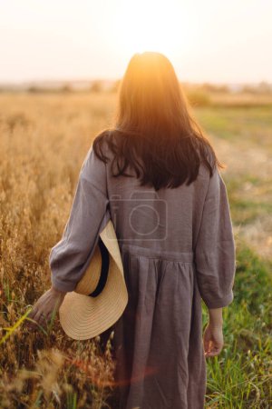 Téléchargez les photos : Femme élégante avec chapeau de paille marchant au champ d'avoine dans la lumière du coucher du soleil, vue arrière. Moment de tranquillité atmosphérique. Jeune femme en robe de lin relaxant en soirée campagne d'été, vie rurale lente - en image libre de droit