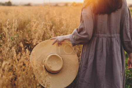 Téléchargez les photos : Femme élégante avec chapeau de paille marchant dans le champ d'avoine dans la lumière du coucher du soleil, vue dégagée. Moment de tranquillité atmosphérique. Jeune femme en robe de lin rustique relaxant en soirée campagne d'été. - en image libre de droit