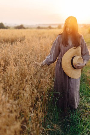 Téléchargez les photos : Femme élégante avec chapeau de paille marchant au champ d'avoine dans la lumière du coucher du soleil. Moment de tranquillité atmosphérique. Jeune femme en robe de lin relaxant en soirée campagne d'été, vie rurale lente - en image libre de droit