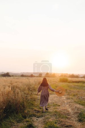Foto de Mujer con estilo con sombrero de paja caminando en el campo de avena en la luz del atardecer, vista trasera. Momento de tranquilidad atmosférica. Mujer joven en vestido de lino relajante en el campo de verano por la noche, vida rural lenta - Imagen libre de derechos