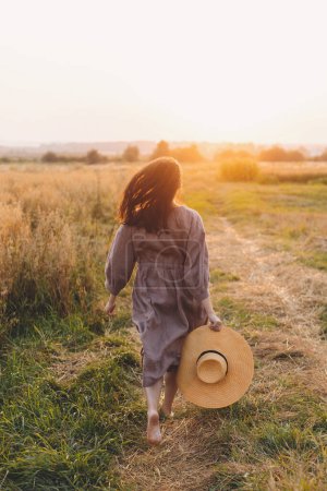 Photo for Stylish woman with straw hat running at oat field in sunset light. Atmospheric happy moment. Young female in rustic linen dress relaxing in evening summer countryside, rural life - Royalty Free Image