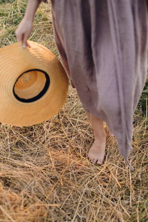 Foto de Mujer con sombrero en la mano caminando descalzo en el campo de paja en la luz del atardecer, vista recortada. Vida rural lenta. Momento de tranquilidad atmosférica. Mujer joven en vestido de lino rústico relajante en el campo de verano - Imagen libre de derechos