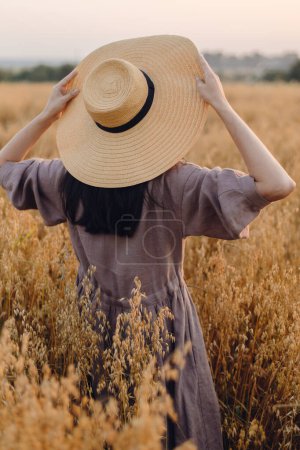 Photo for Stylish woman with straw hat standing in oat field in evening light,  back view. Atmospheric tranquil moment. Young female in rustic linen dress relaxing in summer countryside. Rural slow life - Royalty Free Image