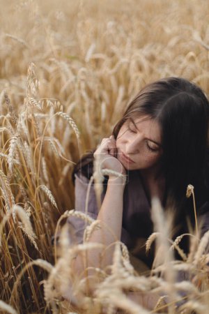 Téléchargez les photos : Femme calme et élégante assise dans un champ de blé en soirée. Moment de tranquillité atmosphérique. Jeune femme en robe de lin rustique relaxant en soirée campagne d'été. Vie rurale lente - en image libre de droit