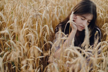Photo for Stylish calm woman sitting in wheat field in evening light. Atmospheric tranquil moment. Young female in rustic linen dress relaxing in evening summer countryside. Rural slow life - Royalty Free Image
