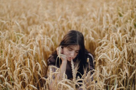 Photo for Wheat stems on background of defocused woman sitting in wheat field in evening. Atmospheric tranquil moment. Ripe ears. Young female relaxing in summer countryside. Rural slow life - Royalty Free Image