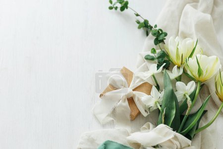 Photo for Beautiful white bouquet with gift box on rustic wooden table flat lay. Happy Mothers day. Stylish spring tulips and daffodils on soft fabric and present. Womens day. Space for text - Royalty Free Image