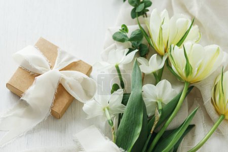 Foto de Feliz Día de las Madres. Hermoso ramo blanco con caja de regalo sobre una mesa de madera rústica. Elegantes tulipanes de primavera y narcisos sobre tela suave y presente. Día de las mujeres - Imagen libre de derechos