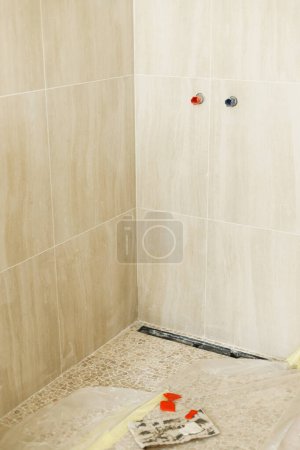 Téléchargez les photos : Tuyau d'eau en tuiles travertin sur le mur et drain de douche dans le sol dans la salle de bain inachevée. Construction d'une maison et rénovation de la maison concept. Canalisations de plomberie - en image libre de droit