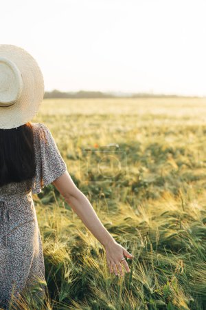 Photo for Beautiful woman in floral dress walking in barley field in sunset light. Atmospheric moment, rustic slow life. Stylish female holding straw hat and enjoying evening summer countryside - Royalty Free Image