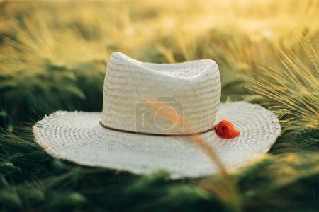 Photo for Summer countryside. Rustic straw hat and red poppy on barley ears in in sunset light in field. Wildflowers and farm hat close up in evening summer meadow. Atmospheric moment - Royalty Free Image