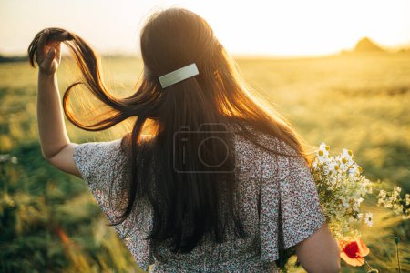 Foto de Hermosa mujer con flores de sauce ramo de pie en el campo de cebada en la luz del atardecer. Elegante mujer relajándose en el campo de verano por la noche y recogiendo flores. Momento de tranquilidad atmosférica - Imagen libre de derechos