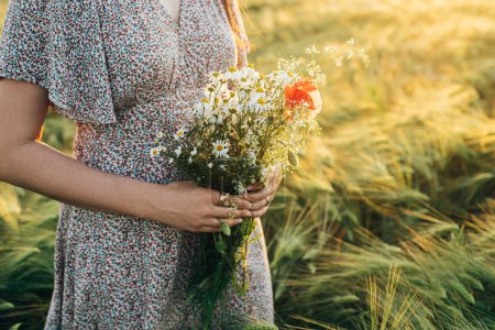 Téléchargez les photos : Beau bouquet de fleurs sauvages dans les mains de la femme près de la lumière du coucher du soleil dans le champ d'orge. Femme élégante relaxant en soirée campagne d'été et cueillette de fleurs. Moment de tranquillité atmosphérique - en image libre de droit