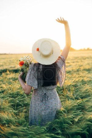 Téléchargez les photos : Belle femme avec des fleurs sauvages profitant du coucher de soleil dans le champ d'orge. Moment tranquille atmosphérique, vie lente rustique. Élégante femelle cueillette de fleurs et de détente en soirée campagne d'été - en image libre de droit
