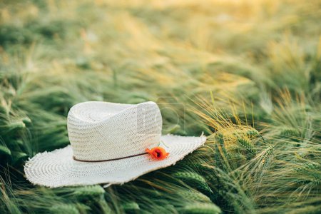 Photo for Rustic straw hat and red poppy on barley ears in evening field close up. Wildflowers and farm hat in summer countryside. Atmospheric moment in evening meadow - Royalty Free Image