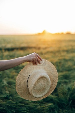 Foto de Mujer sosteniendo sombrero de paja en la luz del atardecer en el fondo del campo de cebada. Elegante mujer relajante en el campo de verano por la noche. Momento tranquilo atmosférico, vida lenta rústica - Imagen libre de derechos