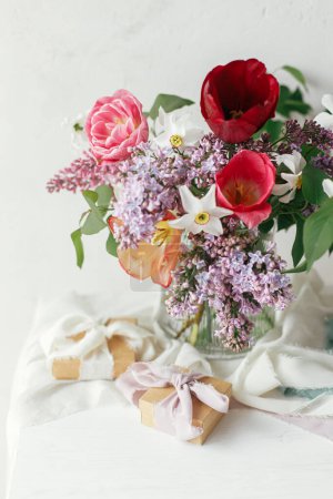 Téléchargez les photos : Bouquet élégant avec coffret cadeau sur table rustique en bois. Bonne fête des mères. Lilas printanier, tulipes, jonquilles arrangement avec présent sur tissu doux dans la salle rustique moderne. Joyeuse journée des femmes - en image libre de droit