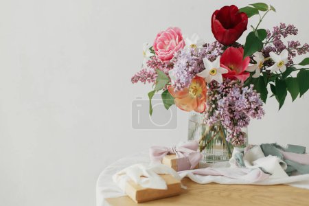 Foto de Feliz Día de las Madres. Primavera lila, tulipanes, narcisos arreglo con cintas presentes y pastel en habitación rústica moderna. Elegante ramo con caja de regalo en una mesa de madera rústica. Feliz día de las mujeres - Imagen libre de derechos