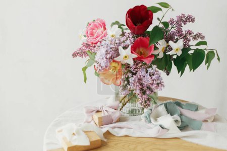 Téléchargez les photos : Bouquet élégant avec coffret cadeau sur table rustique en bois. Bonne fête des mères. Lilas printanier, tulipes, jonquilles arrangement avec présent sur tissu doux dans la salle rustique moderne. Joyeuse journée des femmes - en image libre de droit