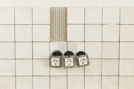 Foto de Elegantes azulejos blancos y toma de corriente se cierran en la pared. Instalación de baldosas cuadradas modernas en caja adhesiva y eléctrica. Renovación de cocina o baño, mejoras para el hogar - Imagen libre de derechos