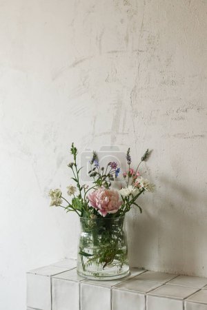 Photo for Stylish bouquet with peony and wildflowers on tile shelf on rustic wall background in modern room. Beautiful flowers in glass vase gathered from garden, summer floral arrangement in home. - Royalty Free Image