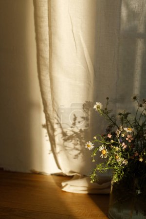Téléchargez les photos : Élégant bouquet de fleurs sauvages dans la lumière du soleil du soir contre la fenêtre dans une pièce rustique. Belles fleurs d'été dans un vase recueilli du jardin, arrangement floral dans la maison de campagne - en image libre de droit