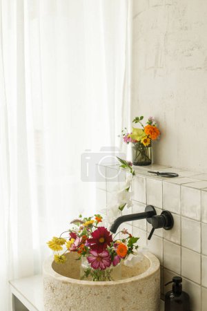 Photo for Beautiful flowers in modern bathroom with stylish sink, black faucet and white tiles. Summer flowers arrangement gathered from garden in modern room in home - Royalty Free Image