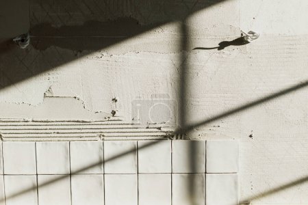 Foto de Elegantes azulejos blancos se cierran en la pared de yeso a la luz del sol. Instalación de baldosas cuadradas modernas en adhesivo. Renovación de cocina o baño, mejoras para el hogar - Imagen libre de derechos