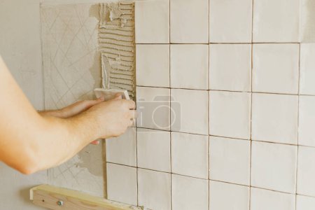 Téléchargez les photos : Travailleur installant des carreaux blancs élégants sur le mur de plâtre à la lumière du soleil. Mains posant carrelage carré moderne sur adhésif fermer. Rénovation cuisine ou salle de bain, rénovation - en image libre de droit
