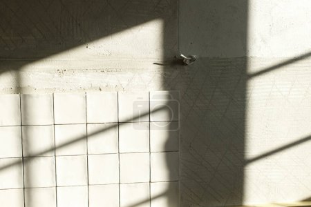 Téléchargez les photos : Les carreaux blancs élégants se ferment sur le mur de plâtre à la lumière du soleil. Carrelage carré moderne posé sur adhésif. Rénovation cuisine ou salle de bain, rénovation - en image libre de droit
