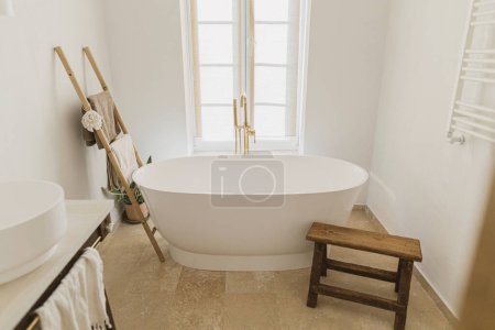 Photo for Stylish bathroom interior.  Modern bathtub with golden faucet from floor, wooden ladder with towels, rustic bench and big window, modern eco bathroom design. - Royalty Free Image