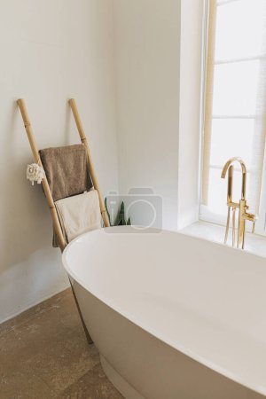 Photo for Stylish boho bathroom design. Modern bathtub with golden faucet from floor, wooden ladder with towels and big window, modern eco bathroom interior. - Royalty Free Image