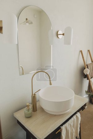 Photo for Modern bathroom interior. Modern ceramic sink with golden faucet on rustic stand with soap and towel on background of wall with mirror and travertine tile. Stylish boho bathroom design. - Royalty Free Image