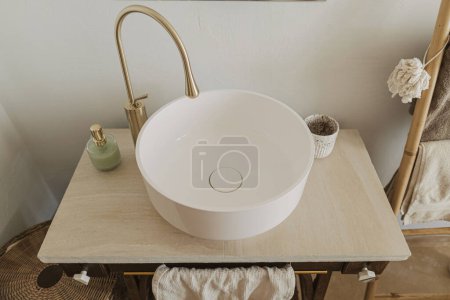 Téléchargez les photos : Design élégant de salle de bains boho. Évier moderne en céramique avec robinet doré sur pied rustique avec savon et serviette sur fond de mur avec miroir et carrelage travertin, intérieur moderne de la salle de bain. - en image libre de droit