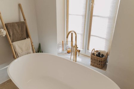 Photo for Stylish boho bathroom design. Modern bathtub with golden faucet from floor, wooden ladder with towels, washing essentials and big window, modern eco bathroom interior. - Royalty Free Image