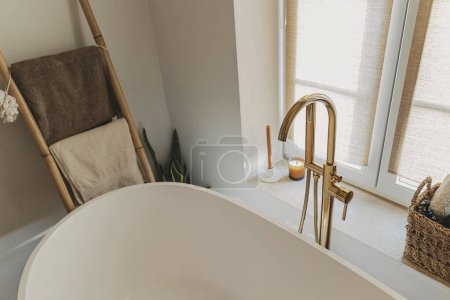 Téléchargez les photos : Design élégant de salle de bains boho. Baignoire moderne avec robinet doré du sol, échelle en bois avec serviettes et grande fenêtre, intérieur moderne éco salle de bain. - en image libre de droit
