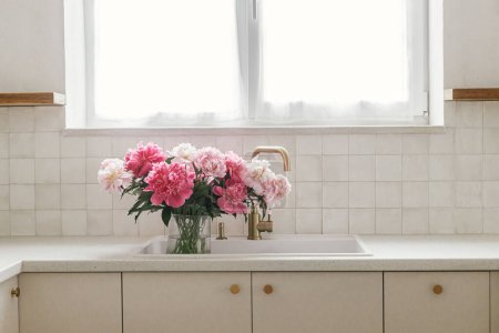 Téléchargez les photos : Belles pivoines dans un vase en évier sur fond de robinet en laiton et fenêtre dans une nouvelle maison scandinave. Fleurs pivoine rose dans la cuisine moderne intérieur, arrangement floral d'été - en image libre de droit